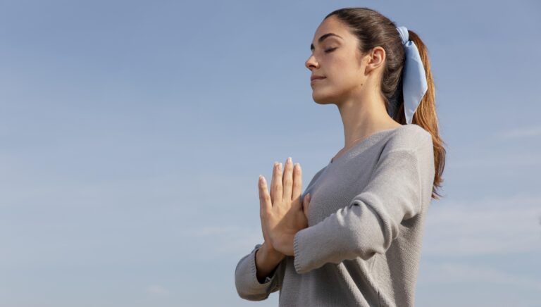 Mujer joven practicando meditación con las manos en posición de oración y los ojos cerrados bajo un cielo despejado.