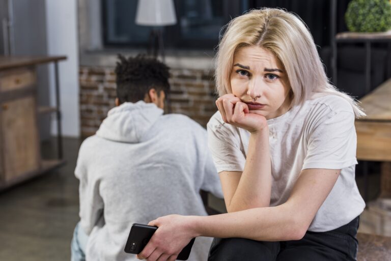 Mujer joven con expresión triste y pensativa sentada junto a su pareja, quien le da la espalda, representando el desapego emocional.